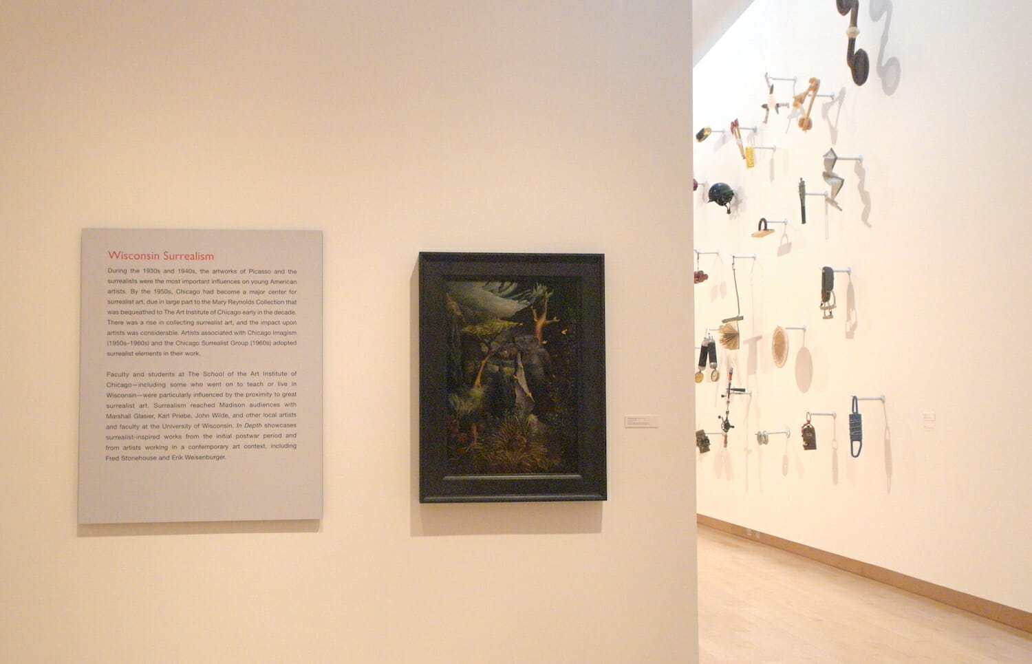 installation view of "In Depth," featuring a framed painting and exhibition text on a wall and another artwork consisting of various objects attached to a wall