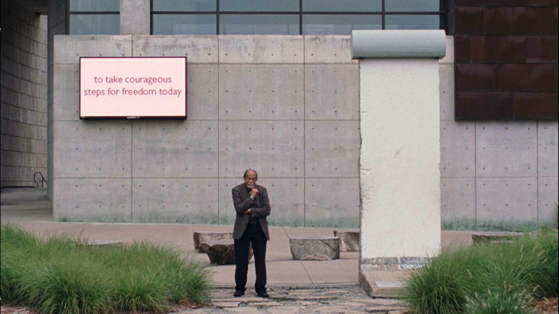 film still showing an older person wearing trousers and a suit blazer standing in front of a building, with a television screen on the exterior that reads "to take courageous steps for freedom today"