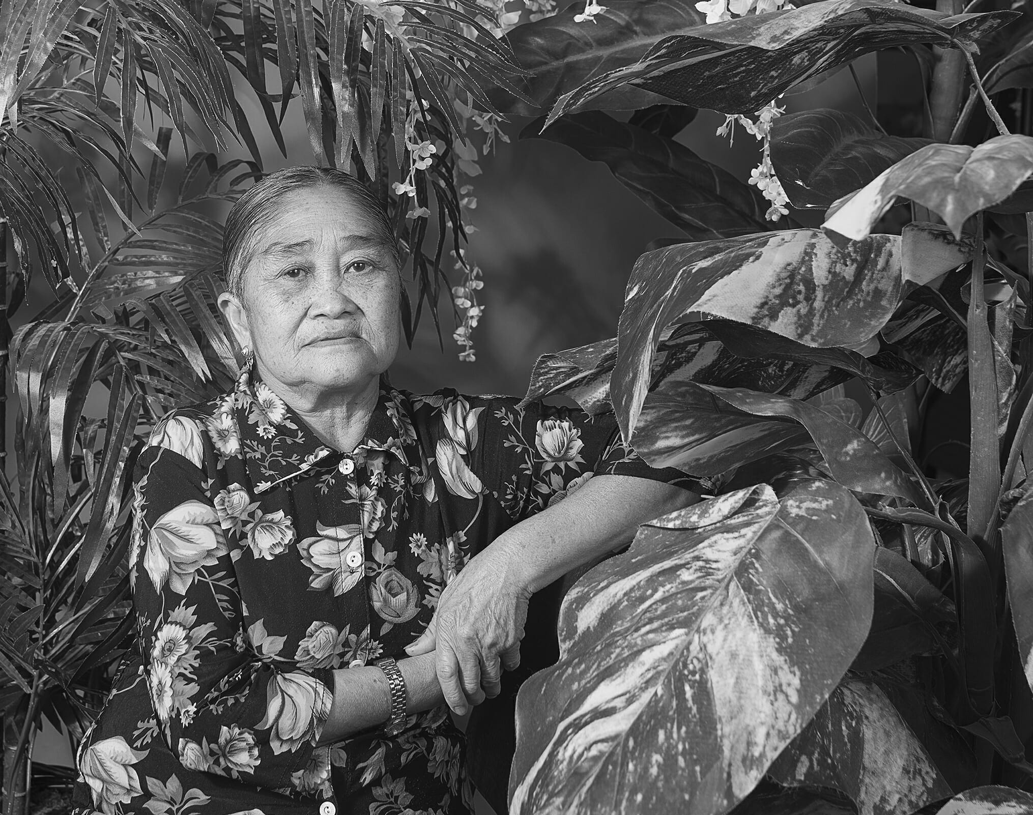 older person wearing a floral print button-up shirt, resting their arm against a surface, in a leafy, vegetal, floral setting