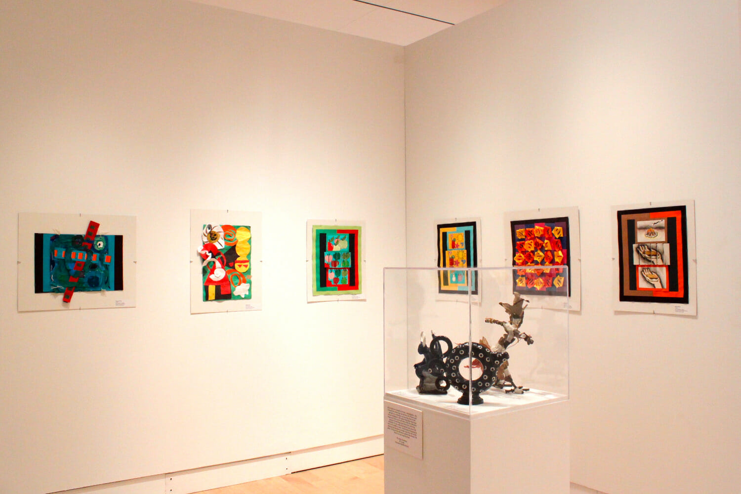 gallery installation view of Young at Art featuring rows of artworks on display across a wall and a vitrine display case with additional sculptures