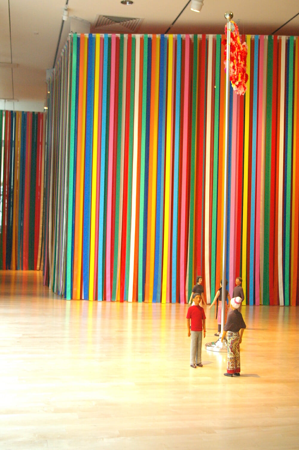 installation view featuring long colorful plastic ribbons hanging from ceiling and touching the floor, with small sculptures of people surrounding a flag pole
