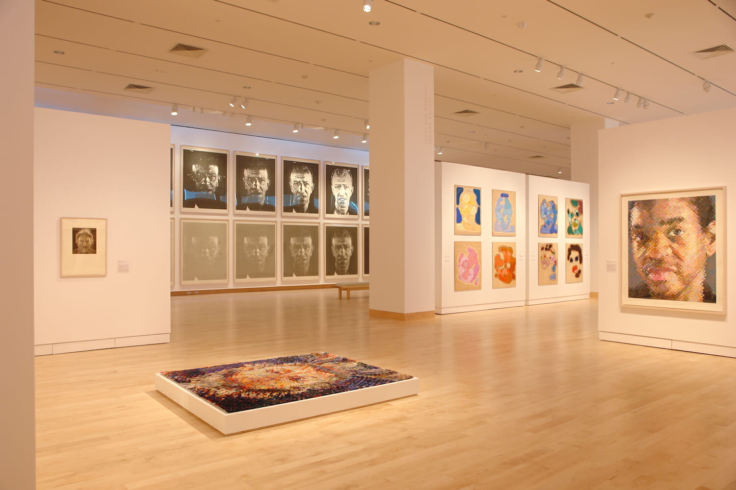 installation view of the Chuck Close Prints exhibition, showing rows of framed artworks displayed on walls and a large artwork in a thick platform or frame on the round