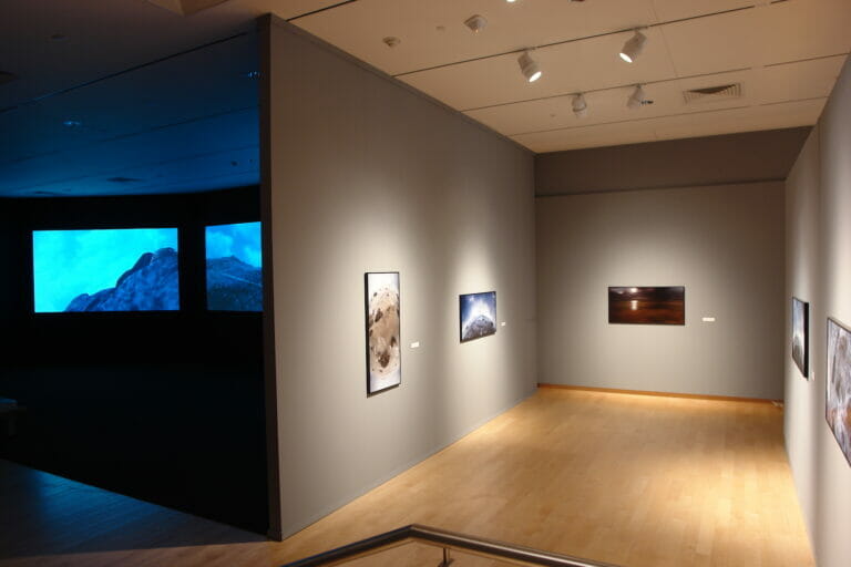 installation view of Cecelia Condit: Within a Stone's Throw showing a dark room playing a video on three screens and a lit gallery with framed photographs on three walls