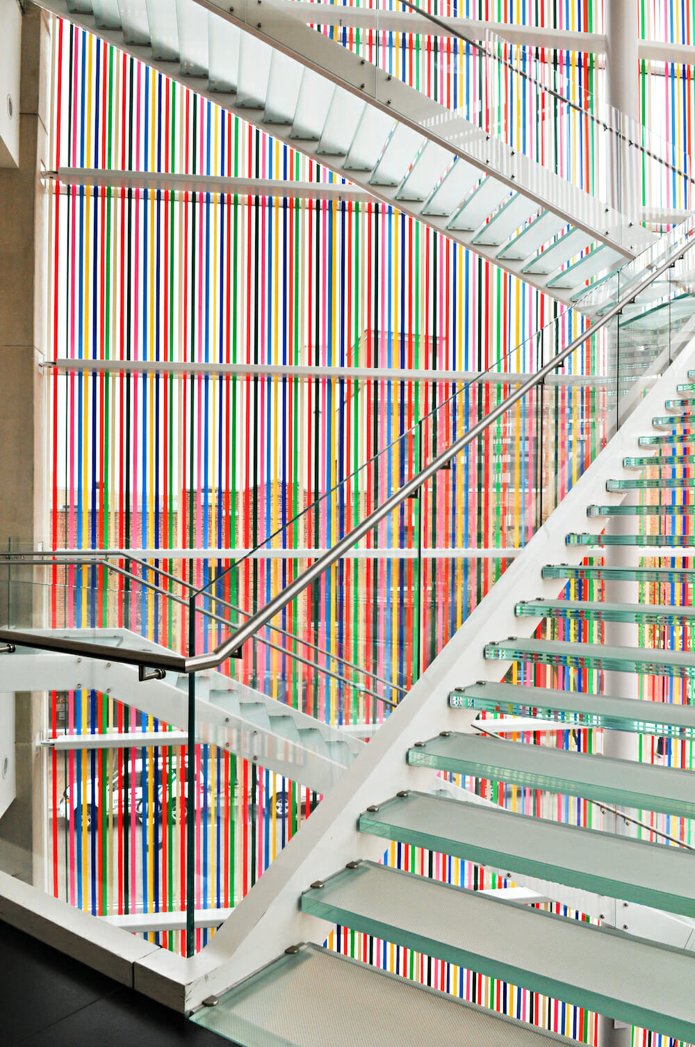installation view of an artwork titled masks, which consists of colorful ribbons hanging down the museum's glass facade, past the glass staircase