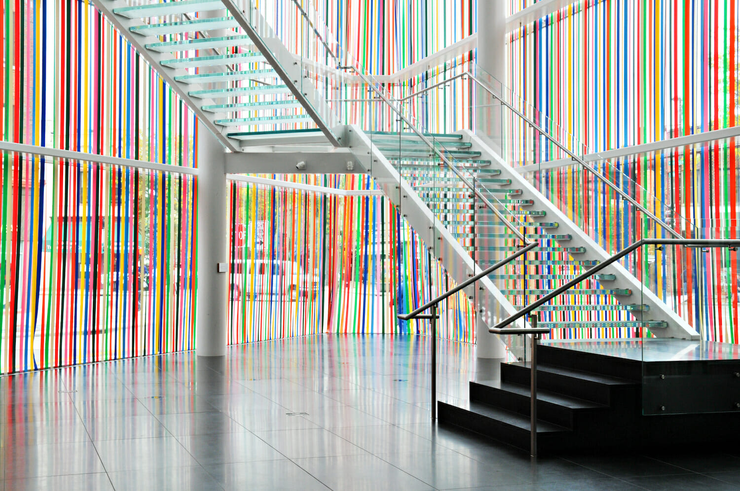 installation view of an artwork titled masks, which consists of colorful ribbons hanging down the museum's glass facade, past the glass staircase