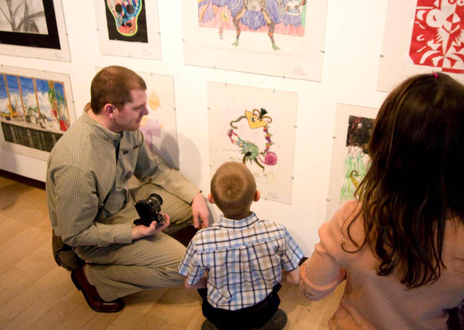 a photographer kneeling or squating down and a child looking at artwork on display on a wall