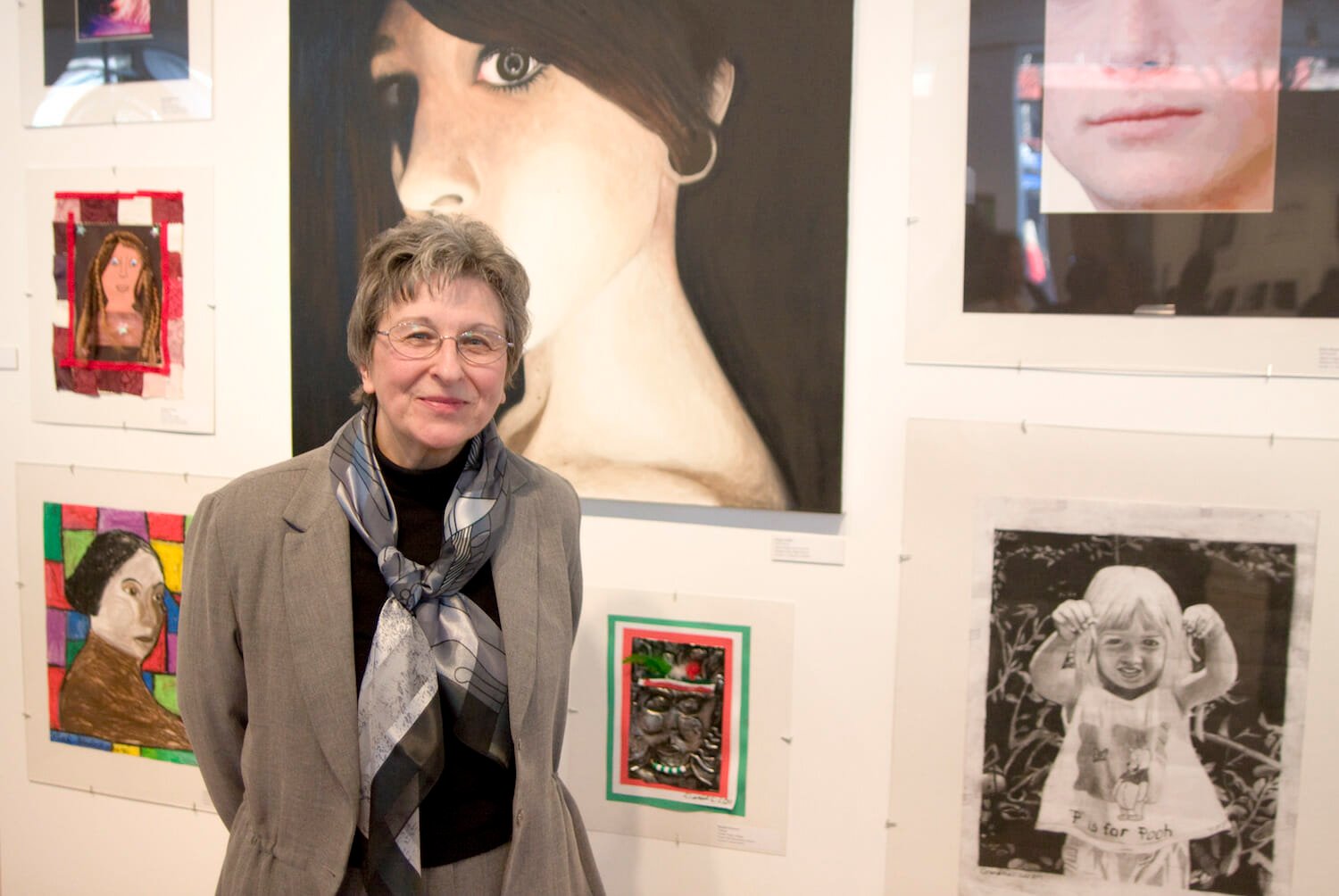 a person with glasses, short hair, a blazer, and a scarf standing in front of artwork made by youth on display on a wall