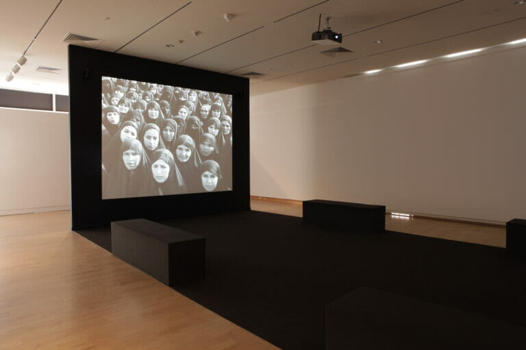 installation view of Shirin Neshat's video titled Rapture, featuring long block benches in front of a black cloth where the video is being projected