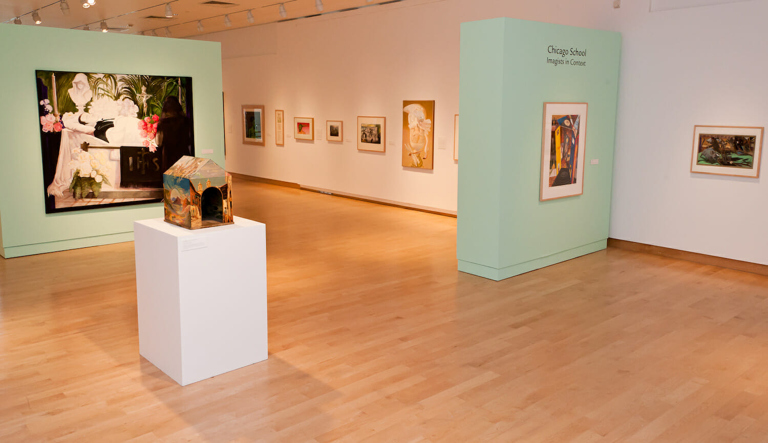 installation view of the exhibition Chicago School featuring a small house made of wood and canvas board on a tall display platform surrounded by artwork hanging vertically across walls