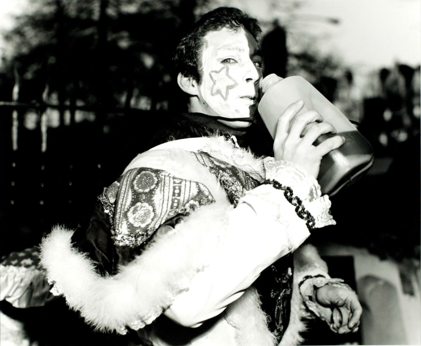 photograph showing a person in costume with face paint, a star painted on their cheek, as they drink out of a plastic bottle
