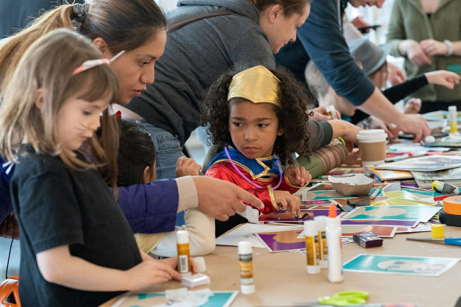 Children are at a table completing art activities. One child is dressed as a superhero, possibly Wonder Woman.