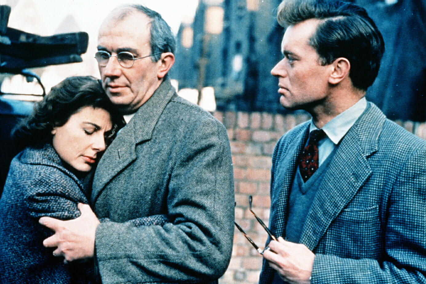 Film still from the 1959 film Sapphire showing a woman hugging and leaning her head on an older man while another man looks at them or to the left of the still. They are all wearing blazers.