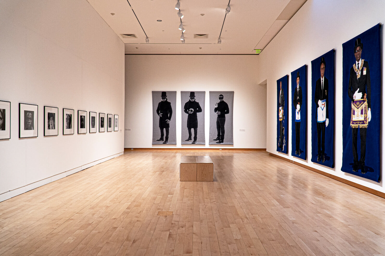 installation view of Faisal Abdu'Allah: DARK MATTER showing three walls covered in hanging artwork and a bench in the center. The left wall has a row of framed photographs. The middle wall has three hanging jacquard tapestries featuring the artist. The right wall shows four of six tapestries featuring members of the Prince Hall Freemasonry.
