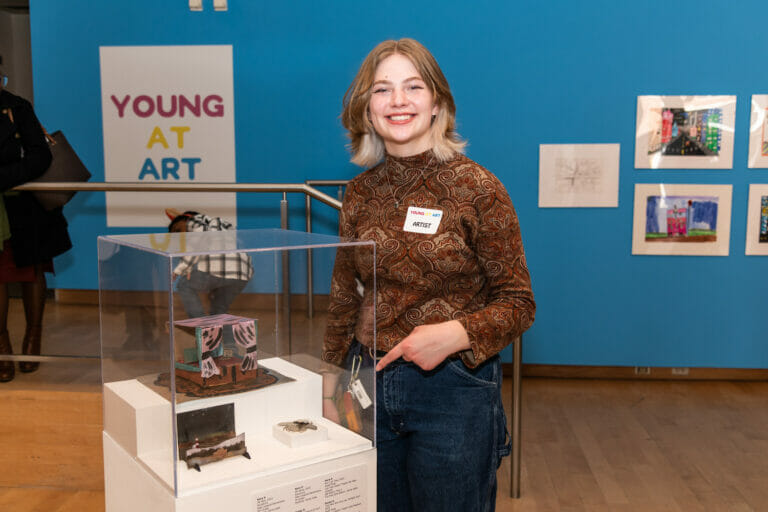 A student artist points at their artwork which is on display in a vitrine display case.