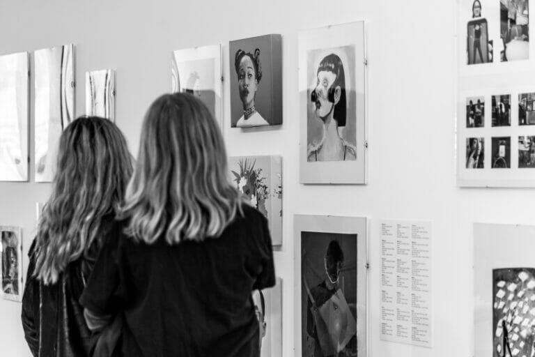 Two people look at artwork which are made by students on display in an exhibition.