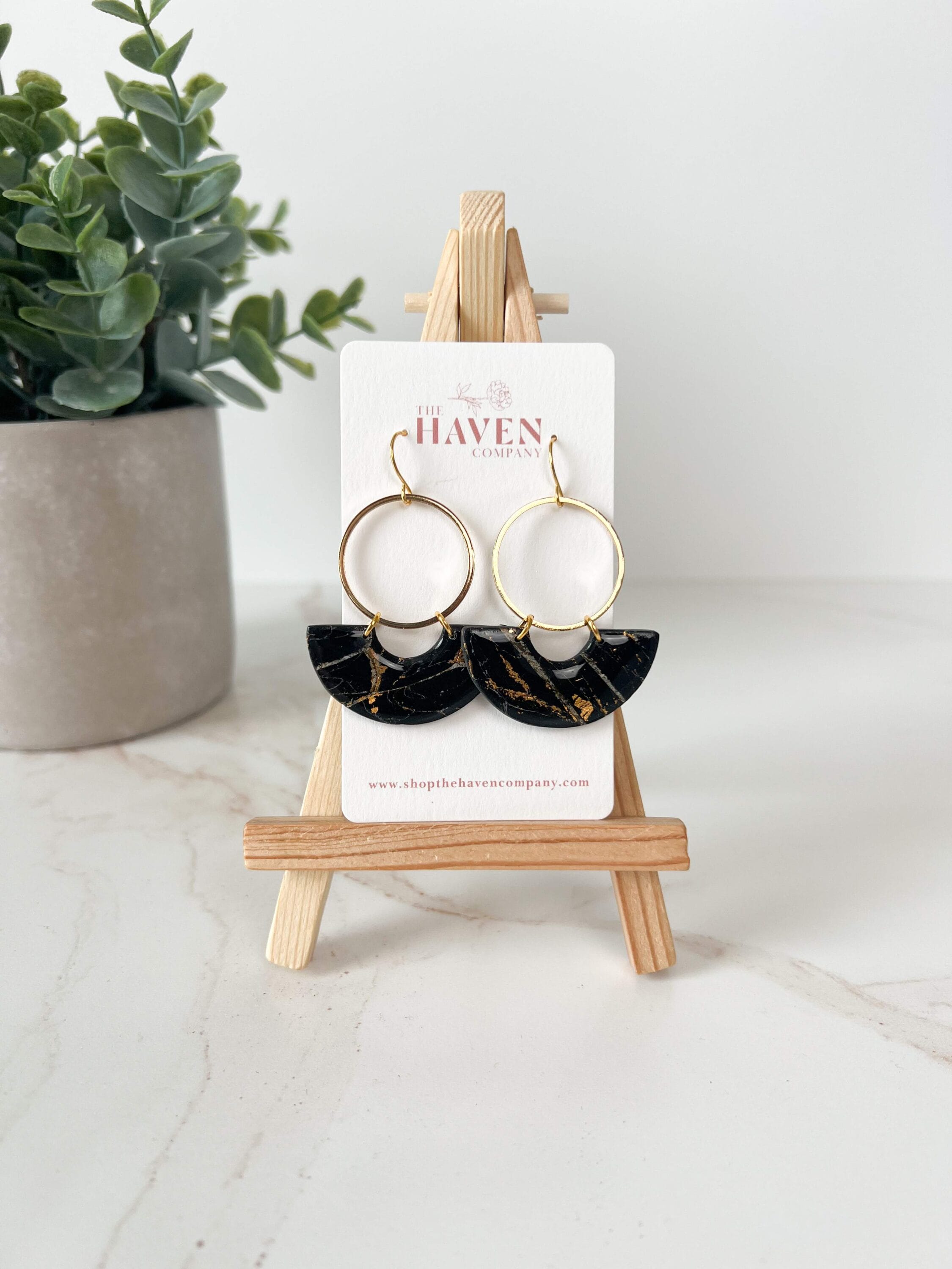 black half circle earrings on a small easel next to a potted plant.