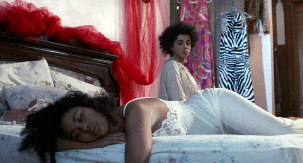Film still from Alma's Rainbow showing one person lying in a bed with their arm hanging off and another person seated behind them on the edge of the bed.