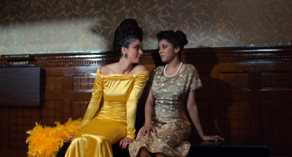 Film still from Alma's Rainbow showing two people seated on a bench while wearing glamorous silk dresses.