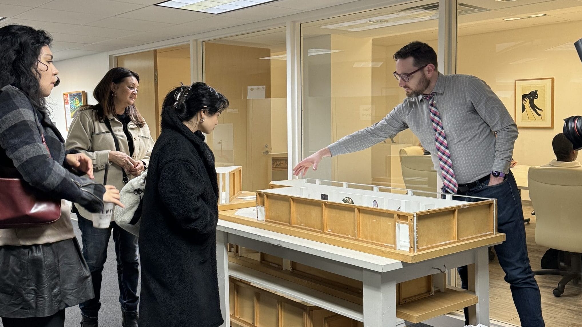 A man wearing a tie pointing at a model of the museum to young college student visitors.