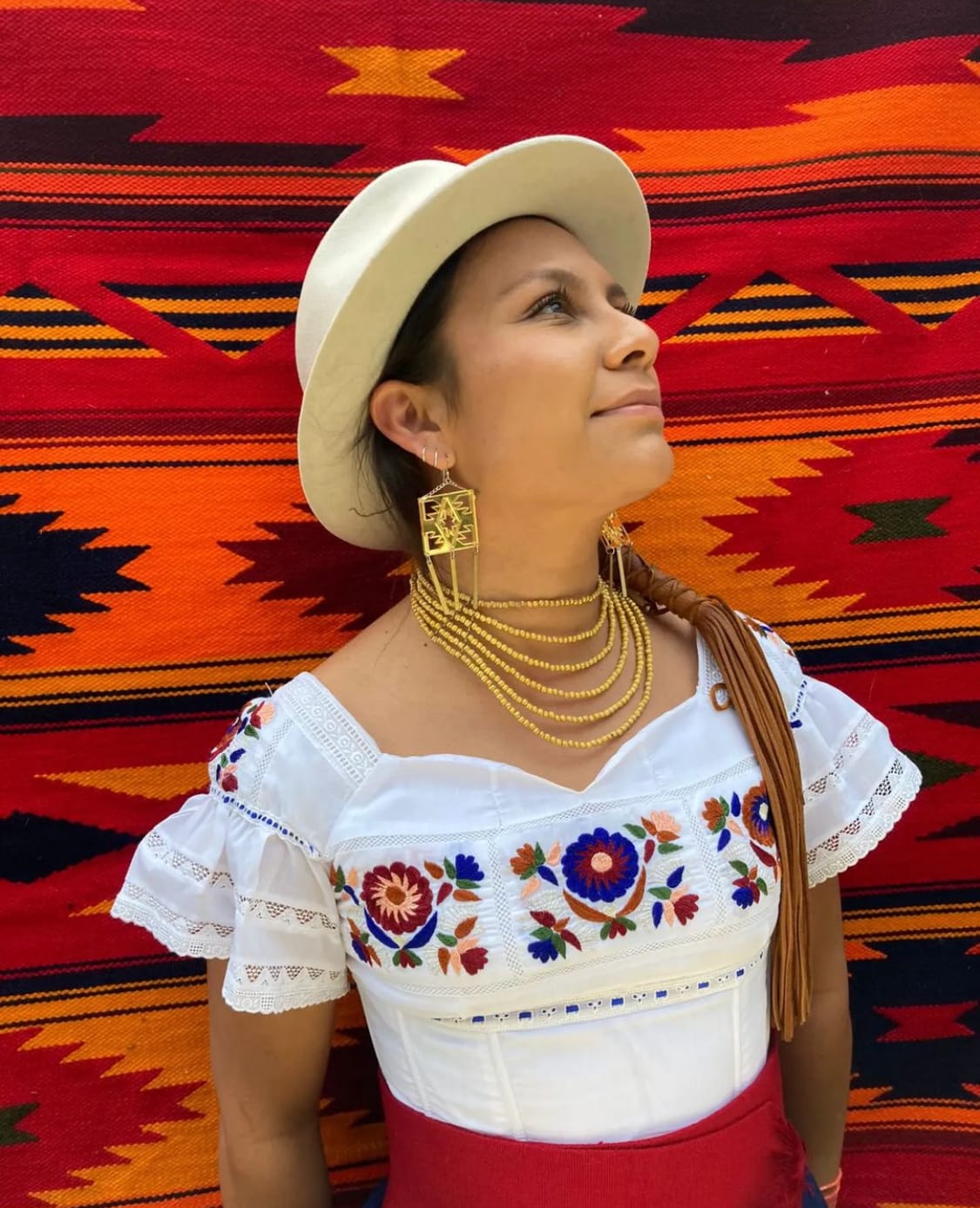 A person looking upwards against a patterned and colorful background while wearing a hat and many necklaces.