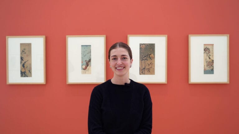 A person smiling in front of art work hung on a pink wall.