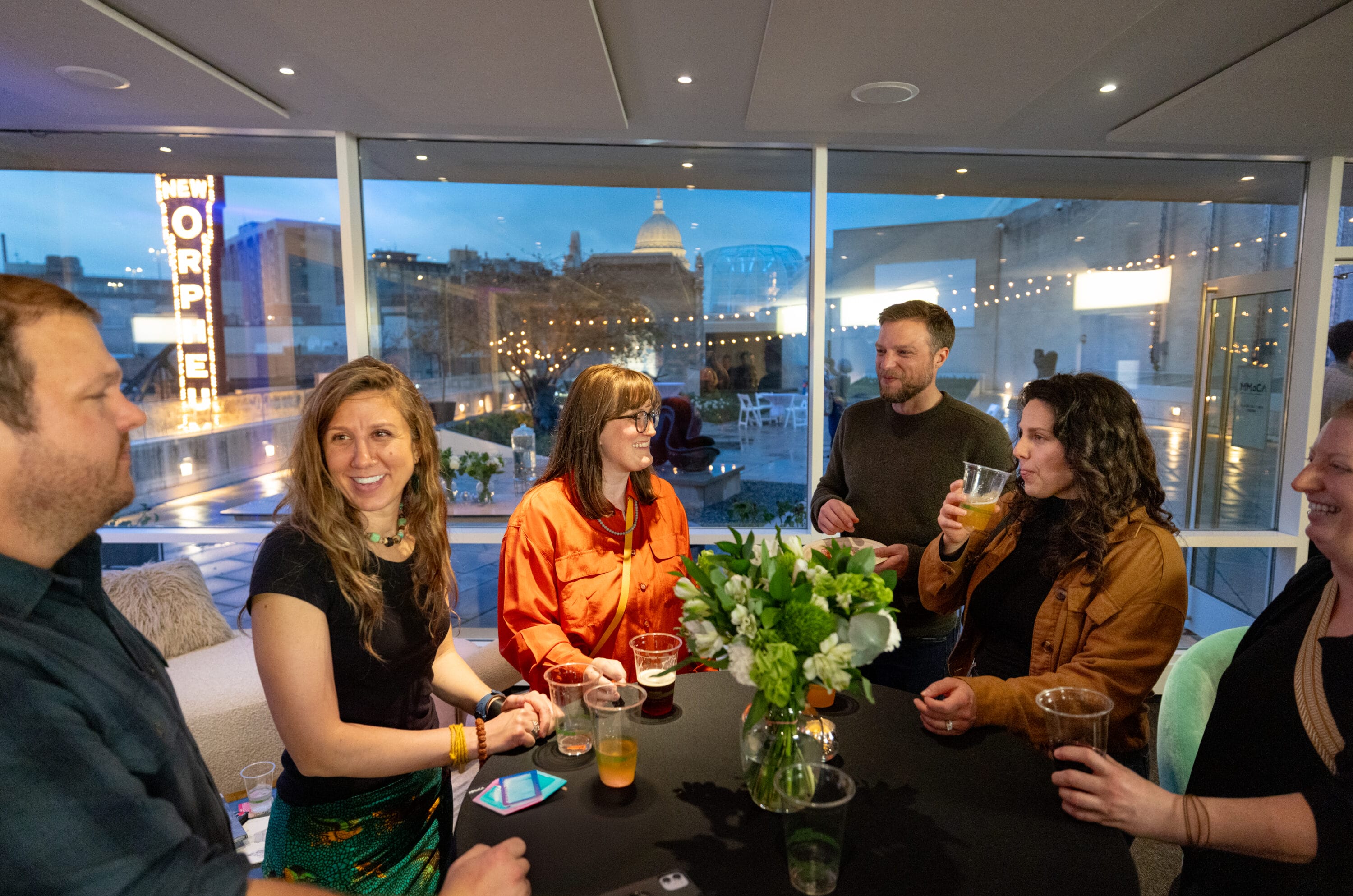 A group of people standing arround a table in front of a window with the madison city skyline in the background.