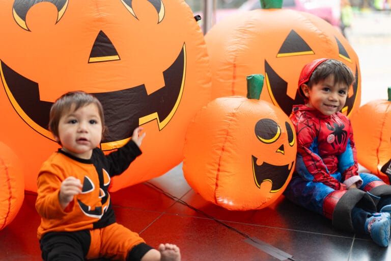 Two kids by pumpkins dressed in Halloween costumes.