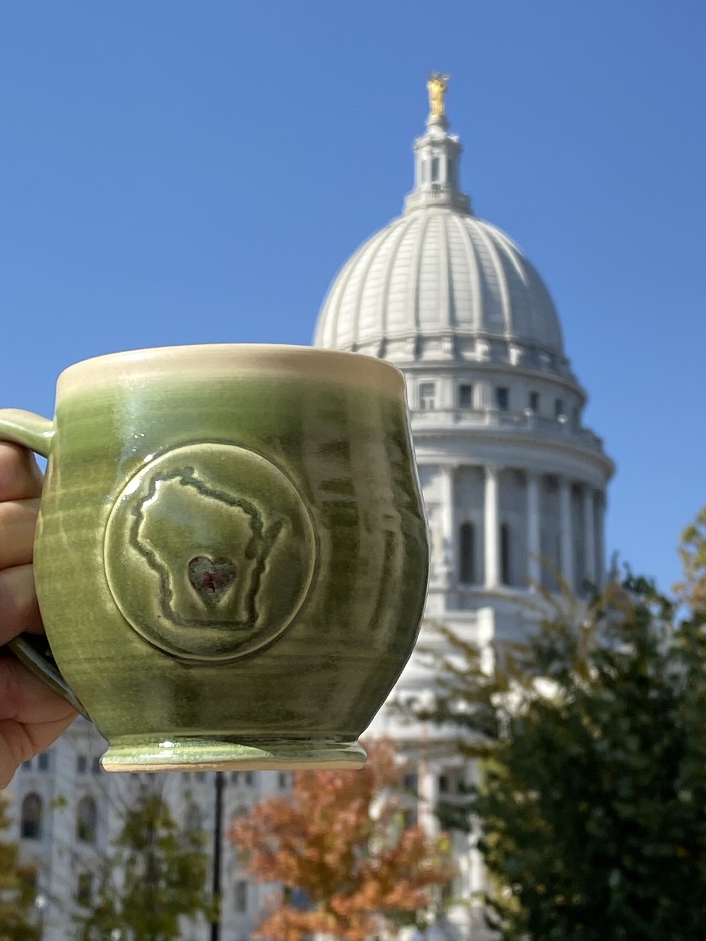 A green mug with the outline of Wisconsin in being held in front of the state capitol building of wisconsin. 