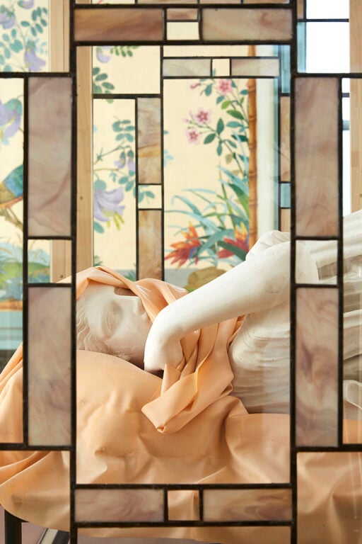 A close up of a marble statue reclining under pink cloth and framed by stained glass.
