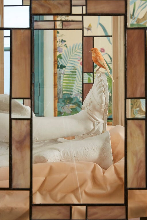 An image of a reclining marble figure with a taxidermied bird on the toe. Framed by stained glass.