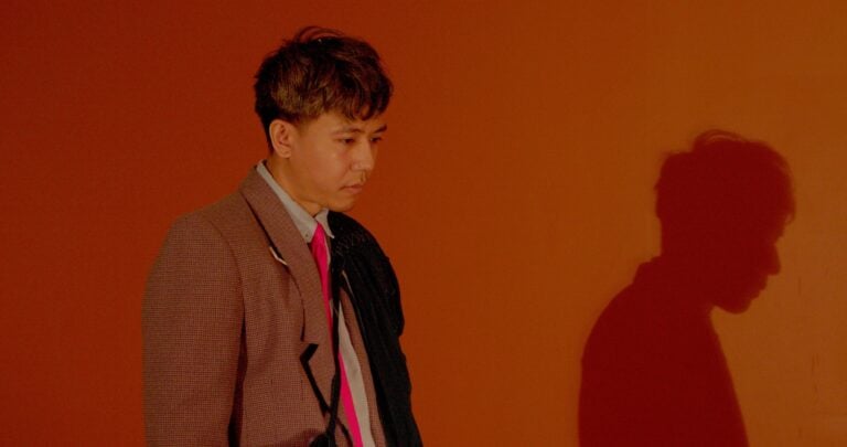 A person in a brown suit against an orange background.