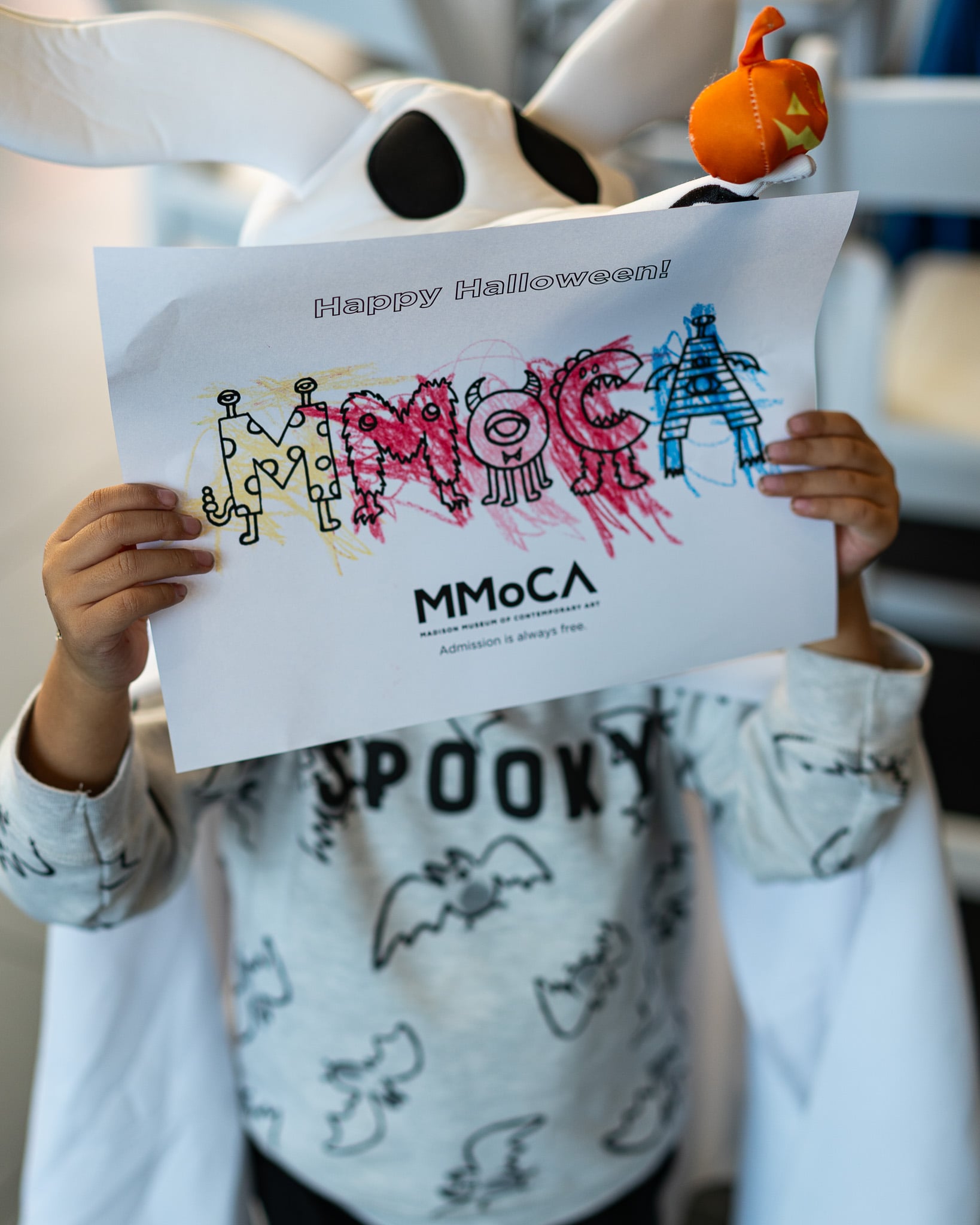 A kid dressed in a ghost dog costume holding a coloring page in front of his face.